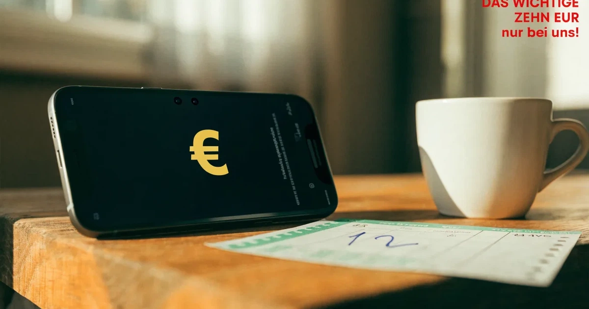 Zwei Euro-Scheine und ein Smartphone liegen auf einem Holztisch neben einer Tasse, im Hintergrund ein unscharfes Fenster.